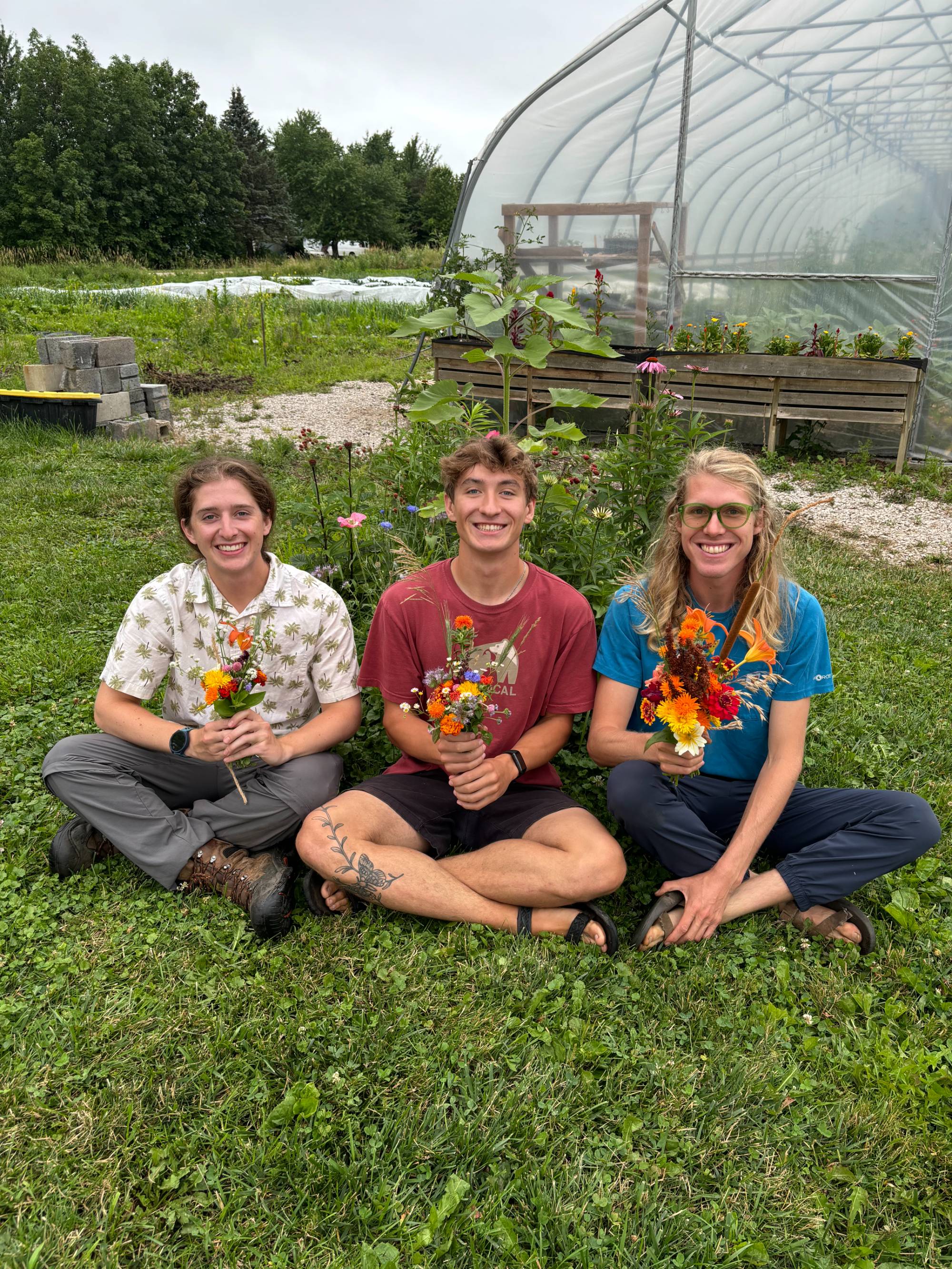 Three students sitting on the ground outside each holding a bouquet of flowers